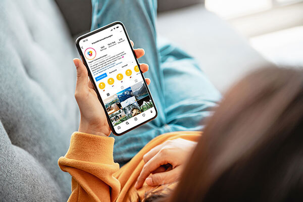 Junge Frau liegt auf dem Sofa und hält ein Handy in der Hand, auf dem der Instagram Account von Traum-Ferienwohnungen zu sehen ist