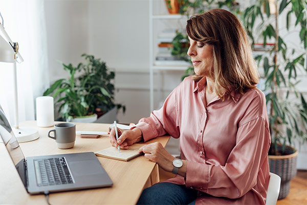 Frau sitzt am Laptop und schreibt einen Text in einem Notizbuch